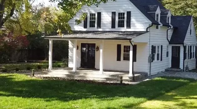 White house with a front porch and dormer windows, surrounded by a green lawn and trees.