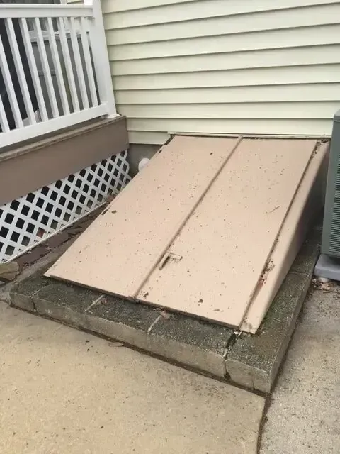 Tan concrete cellar hatch beside a house with beige siding and white railing.