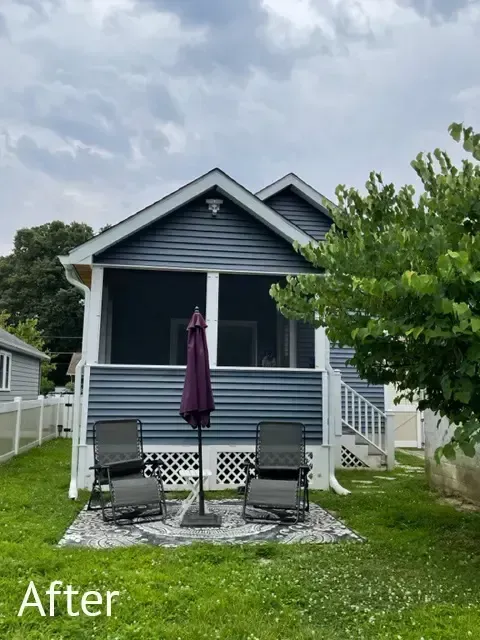 Small blue house with front porch, two chairs, and a closed purple umbrella under a cloudy sky.