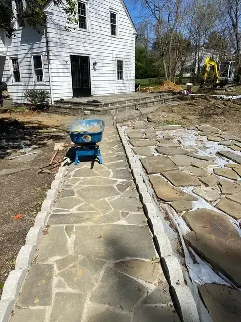 Stone walkway under construction beside a white house, with a blue wheelbarrow and excavator in the yard