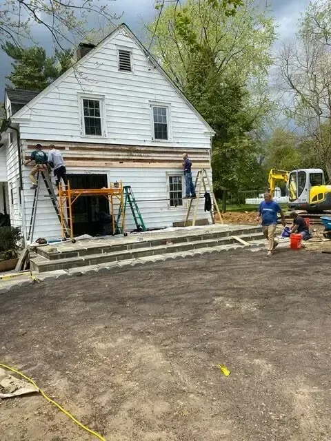 Workers installing siding on a white house exterior with ladders and construction equipment in front yard