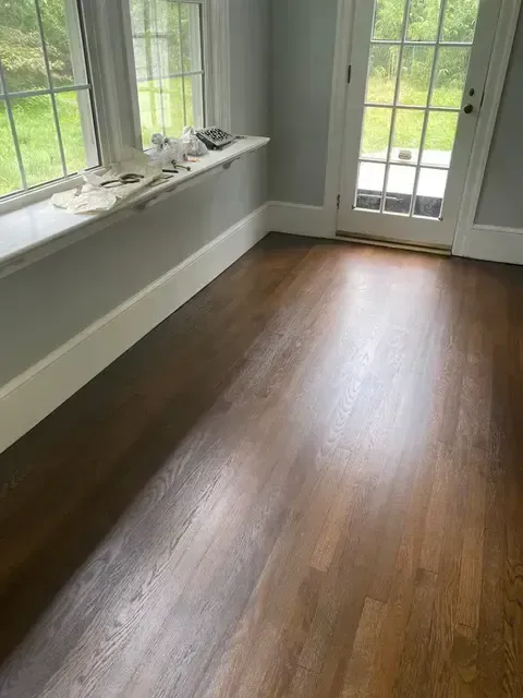Empty room with dark wood floors, light walls, and tall windows opening to a green yard