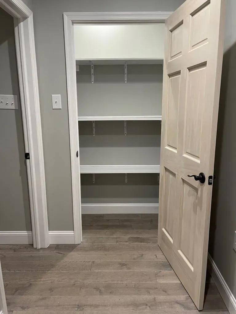 Open closet with white shelves and beige door in a hallway with wood flooring.