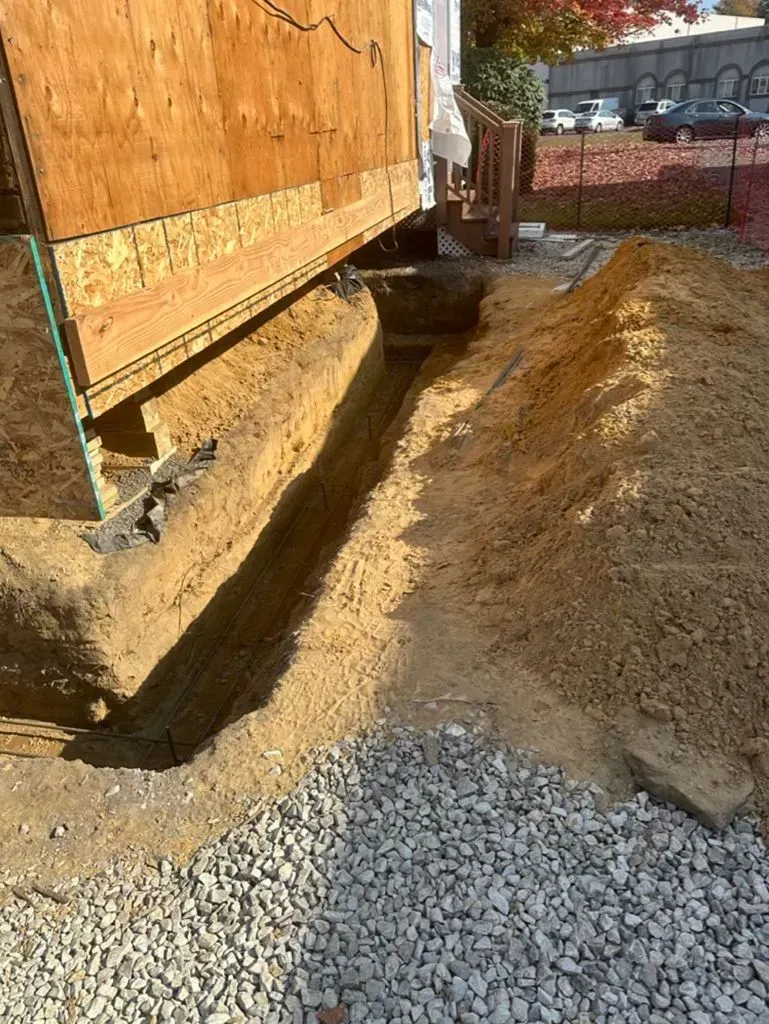 Construction trench along a wooden building beside gravel and dirt piles, with a wheelbarrow in the background