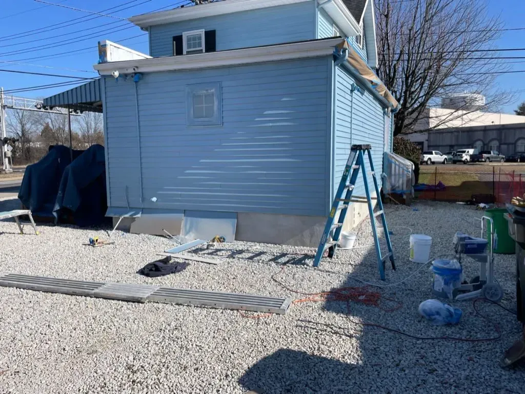 Snow-covered backyard beside a light blue house with a ladder leaning against the wall.