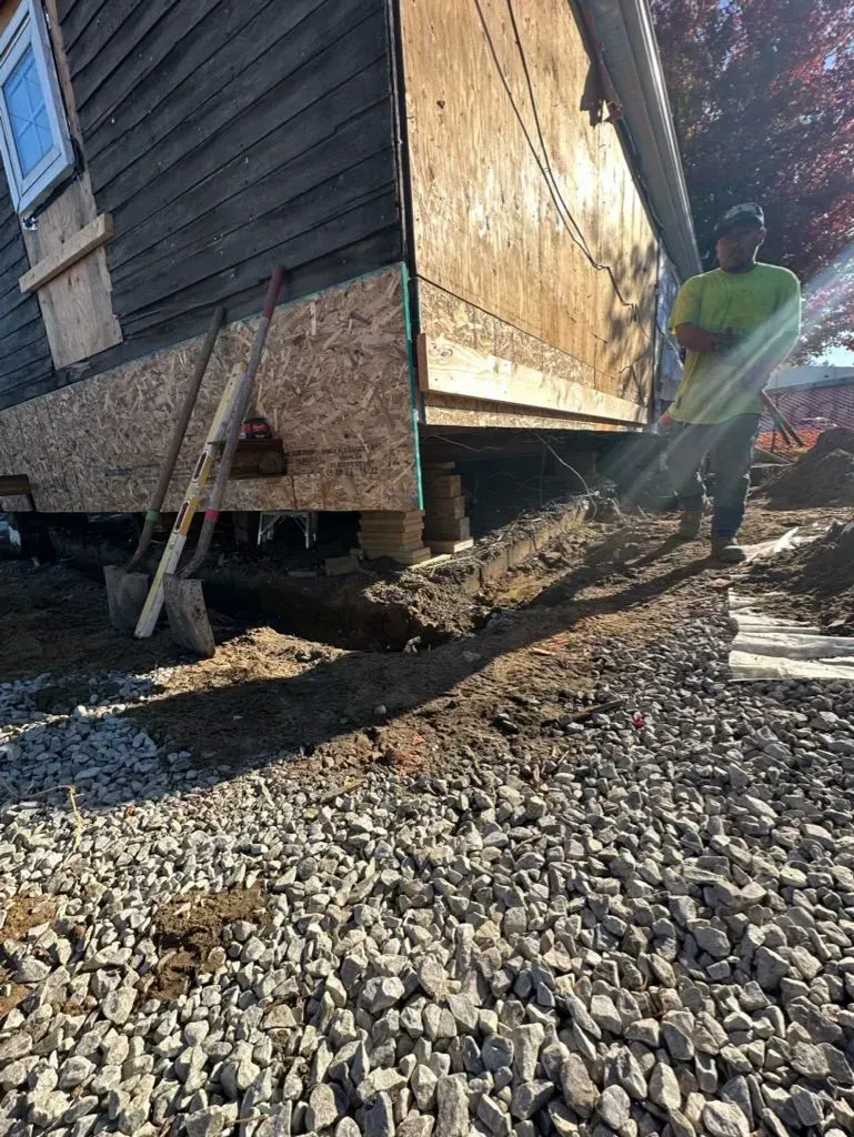 House being lifted on jacks over gravel, with a worker standing nearby in sunlight.