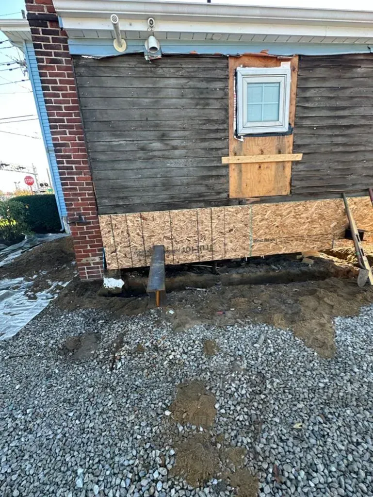 Damaged house wall with exposed wooden siding, a small window, and gravel ground below