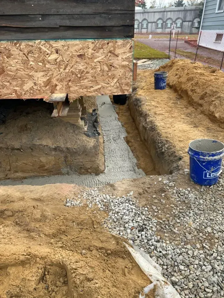 Foundation excavation beside a house, with trench, gravel, and blue buckets in a construction site.