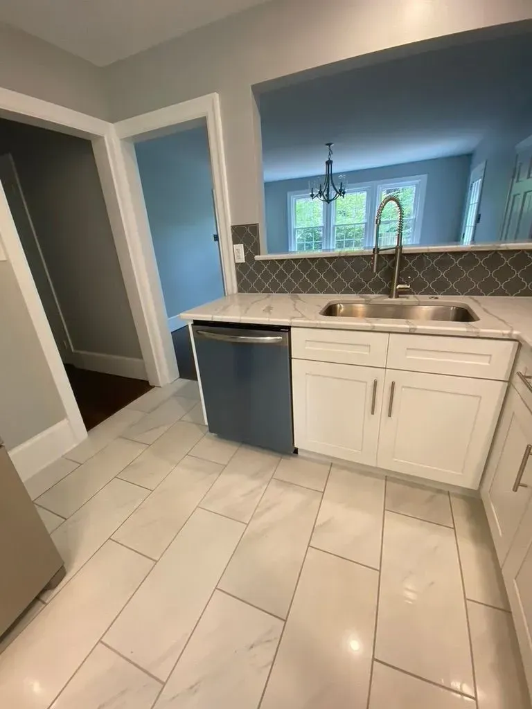 Bright kitchen with white cabinets, tiled floor, and a black dishwasher, looking into a dining area.