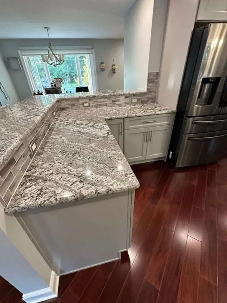Kitchen with granite island, stainless fridge, dark wood floors, and a bright dining area in the background