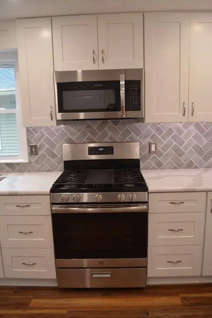 White kitchen with stainless steel stove and microwave beneath white cabinets and gray backsplash.