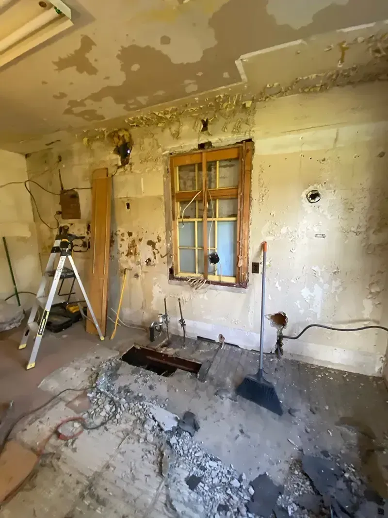 Dilapidated room with peeling walls, debris on floor, ladder, and a small window.