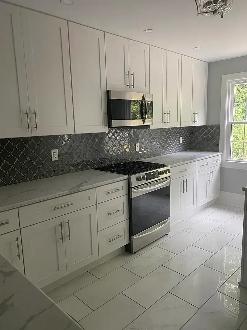 Bright kitchen with white cabinets, dark mosaic backsplash, stainless stove, and large window.
