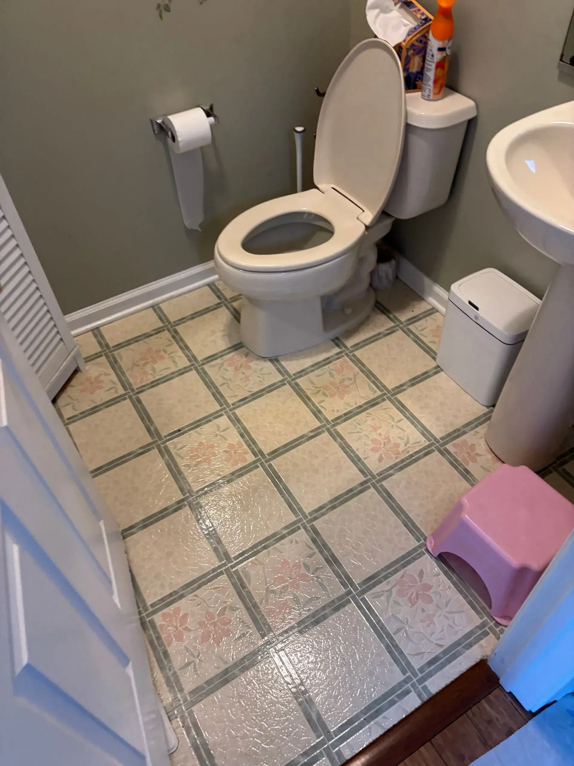 Small bathroom with an open toilet, sink, toilet paper holder, and pink step stool on tiled floor