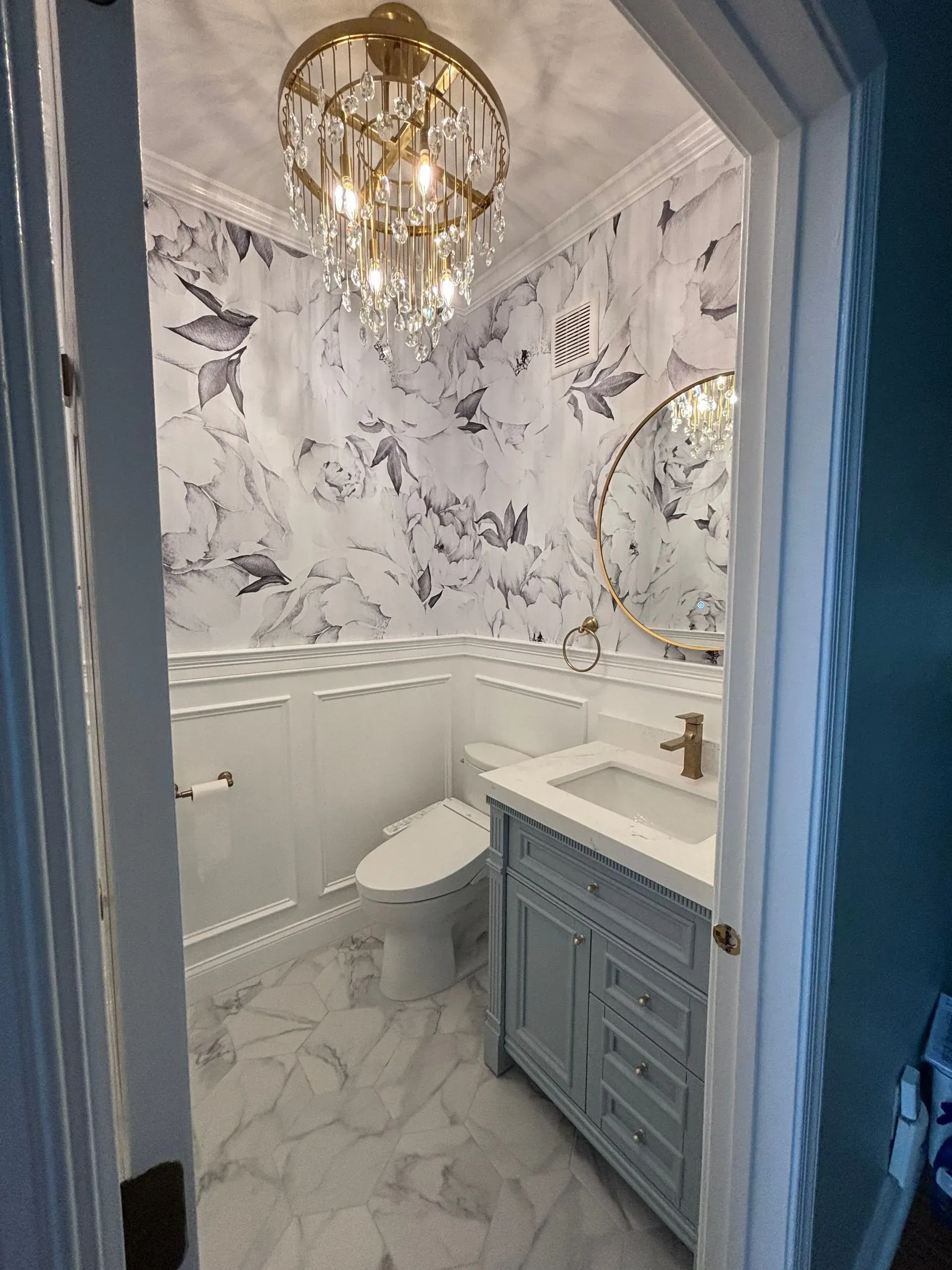 Elegant marble bathroom with chandelier, blue vanity, and toilet, viewed through a doorway
