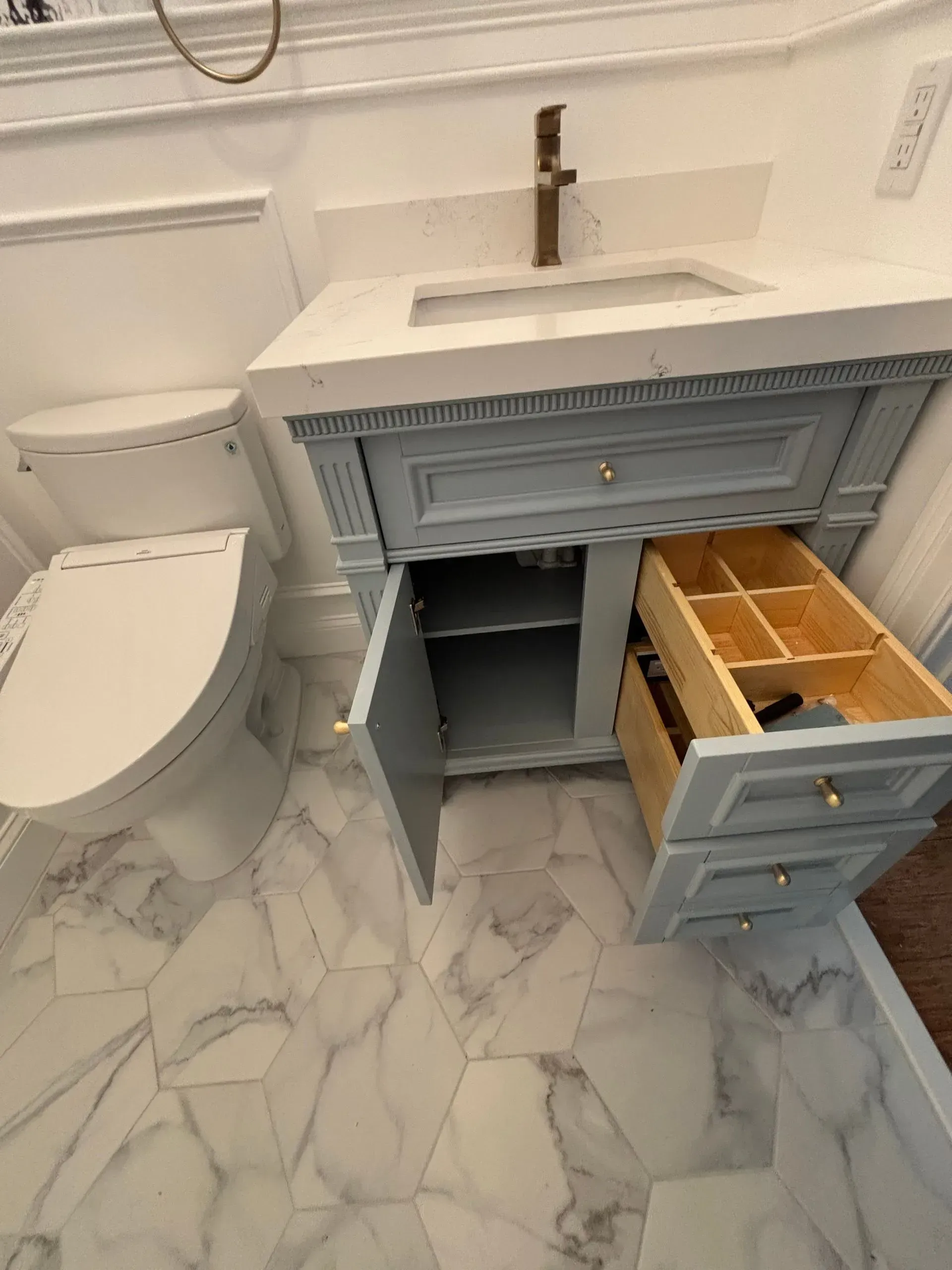 Gray bathroom vanity with open cabinet and a drawer removed, showing storage compartments and a marble floor