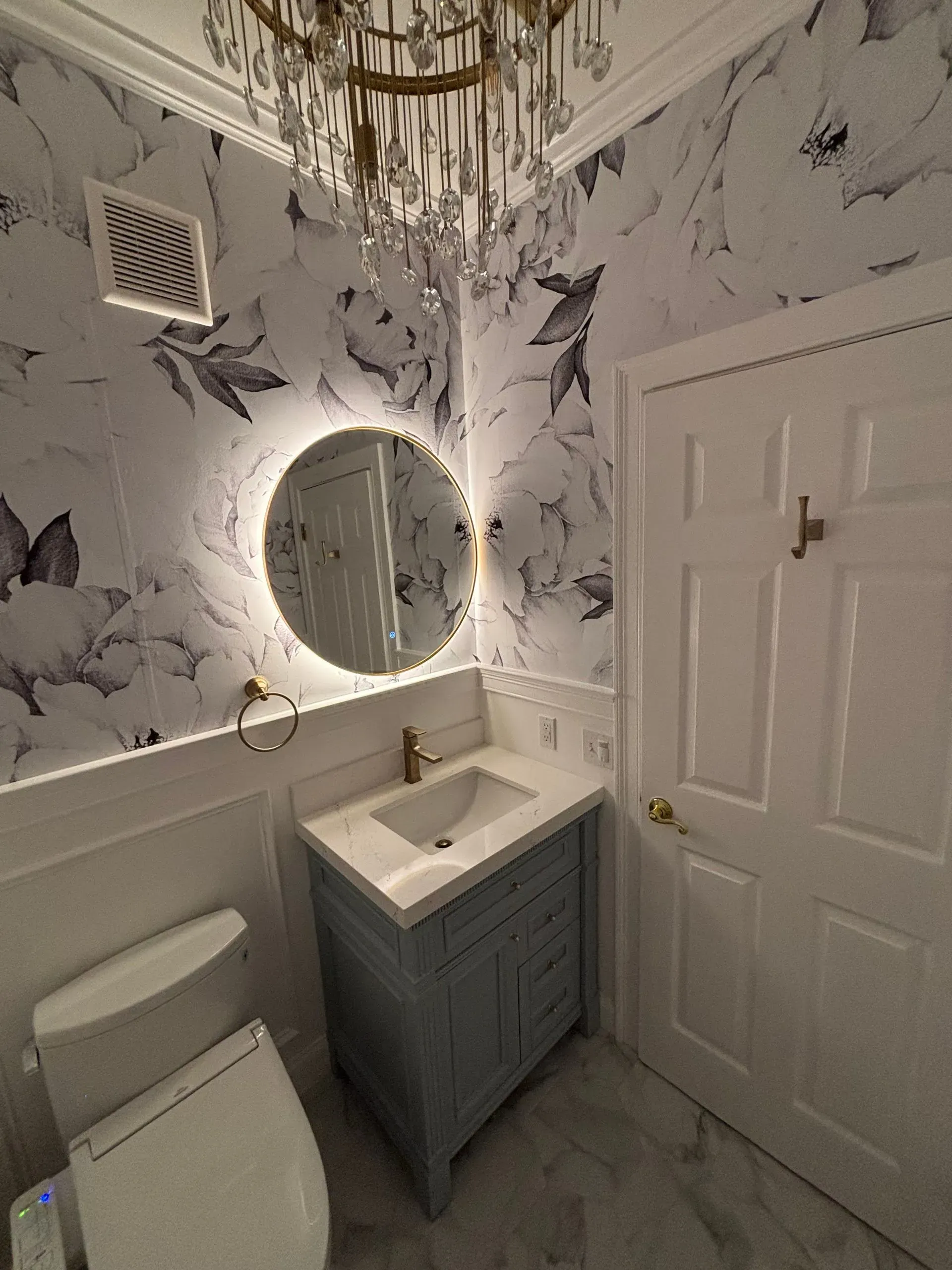 Small bathroom with floral wallpaper, round lit mirror, white vanity, toilet, and white door.