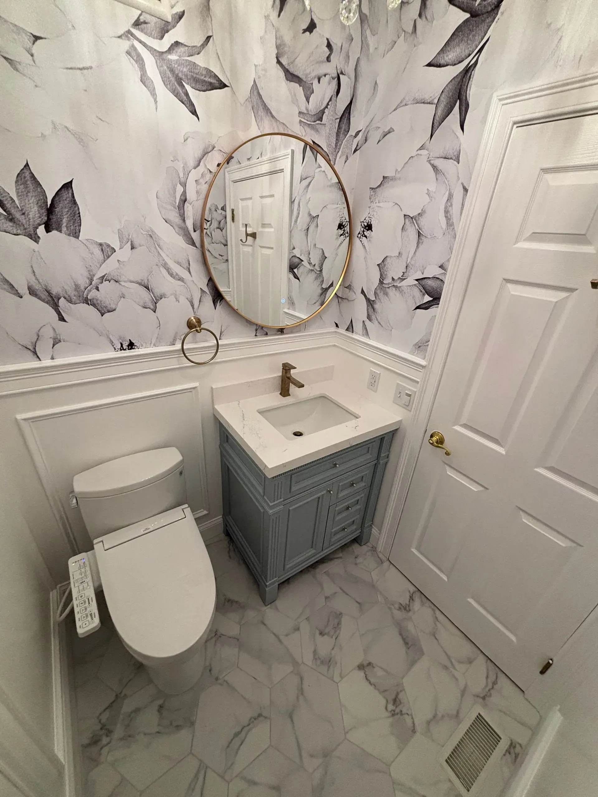 Small bathroom with marble wallpaper, white toilet, patterned vanity, round mirror, and white door