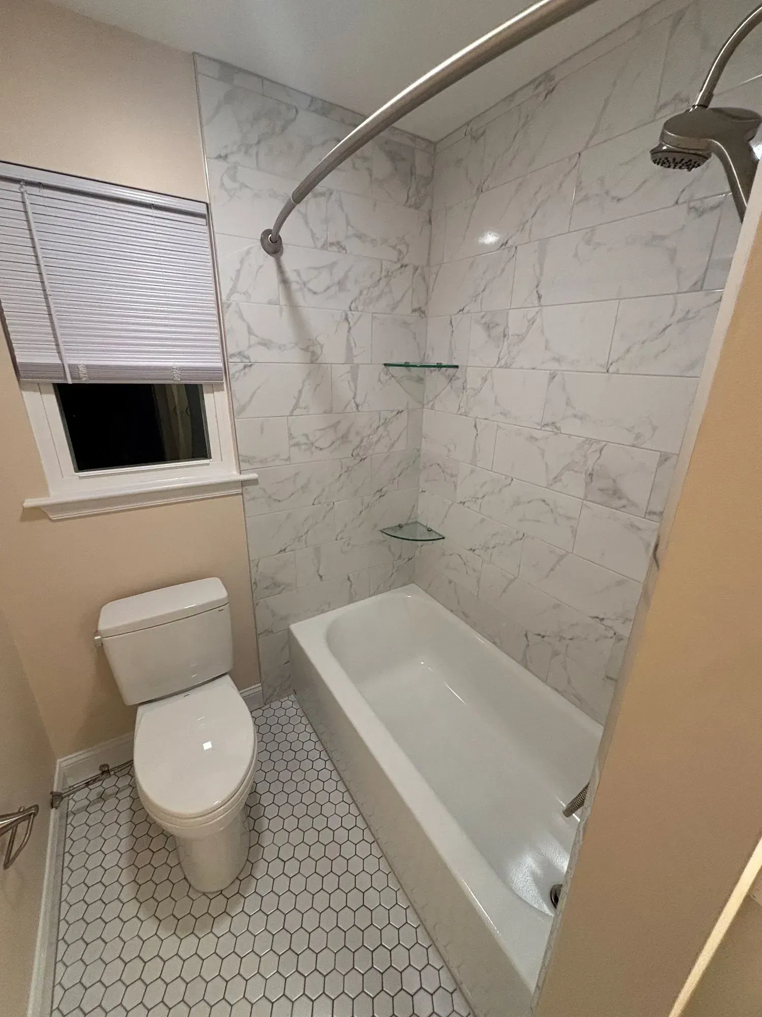 Small bathroom with marble shower wall, bathtub, toilet, and a window with blinds.