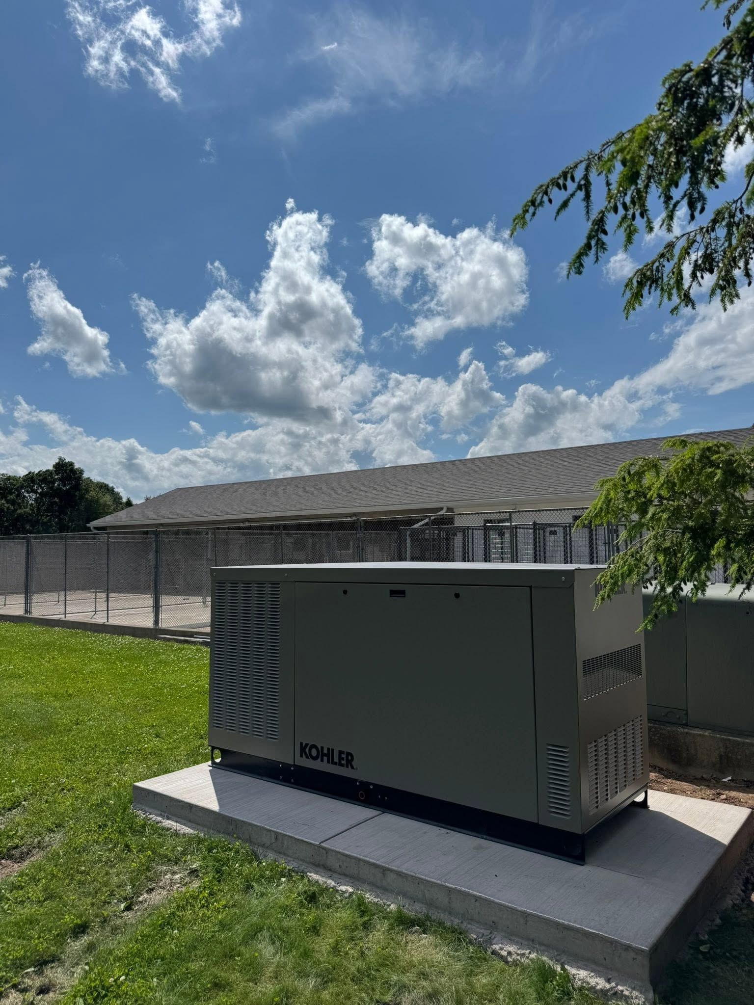 Outdoor electrical generator unit on a concrete pad beside a fenced area under a blue sky with clouds