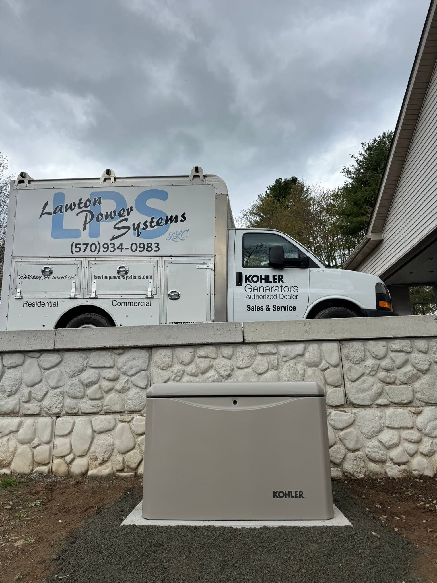 White utility truck on a street behind a stone wall, with a rooftop unit in front and cloudy sky.