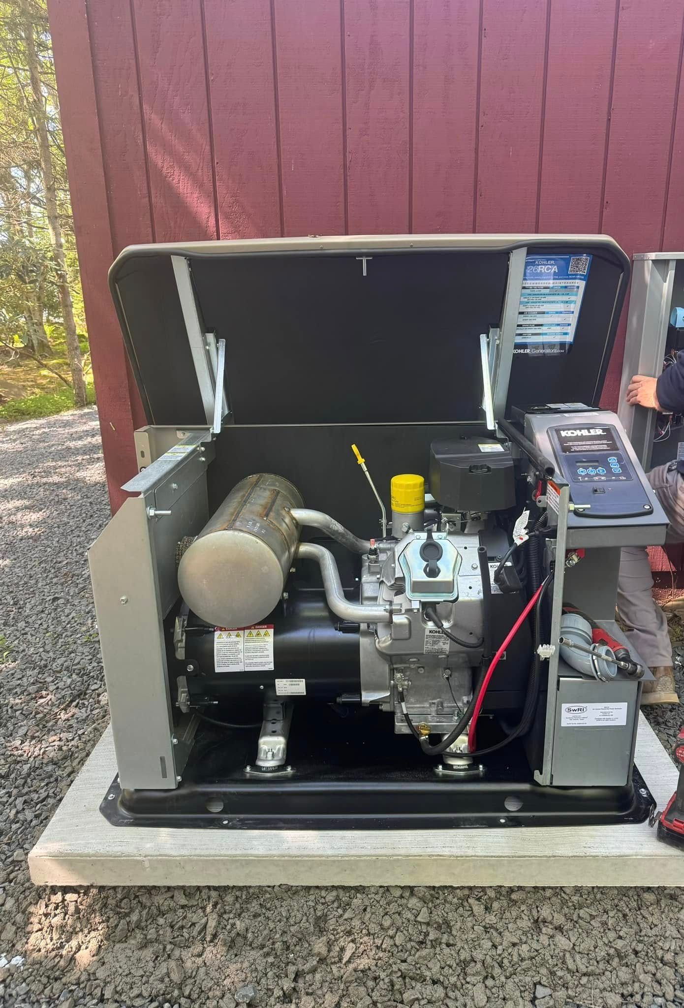 Open generator in a metal enclosure on a concrete pad beside a red wall.