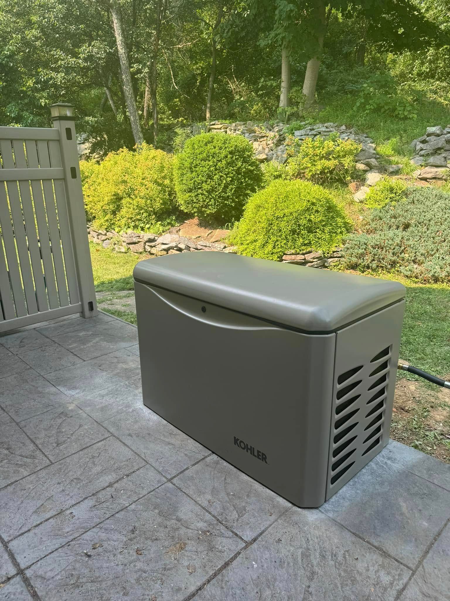 Gray outdoor storage box on a patio beside a gate, with a garden and shrubs in the background