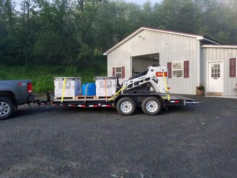 Utility trailer loaded with boxes parked beside a white workshop building on a gravel lot.
