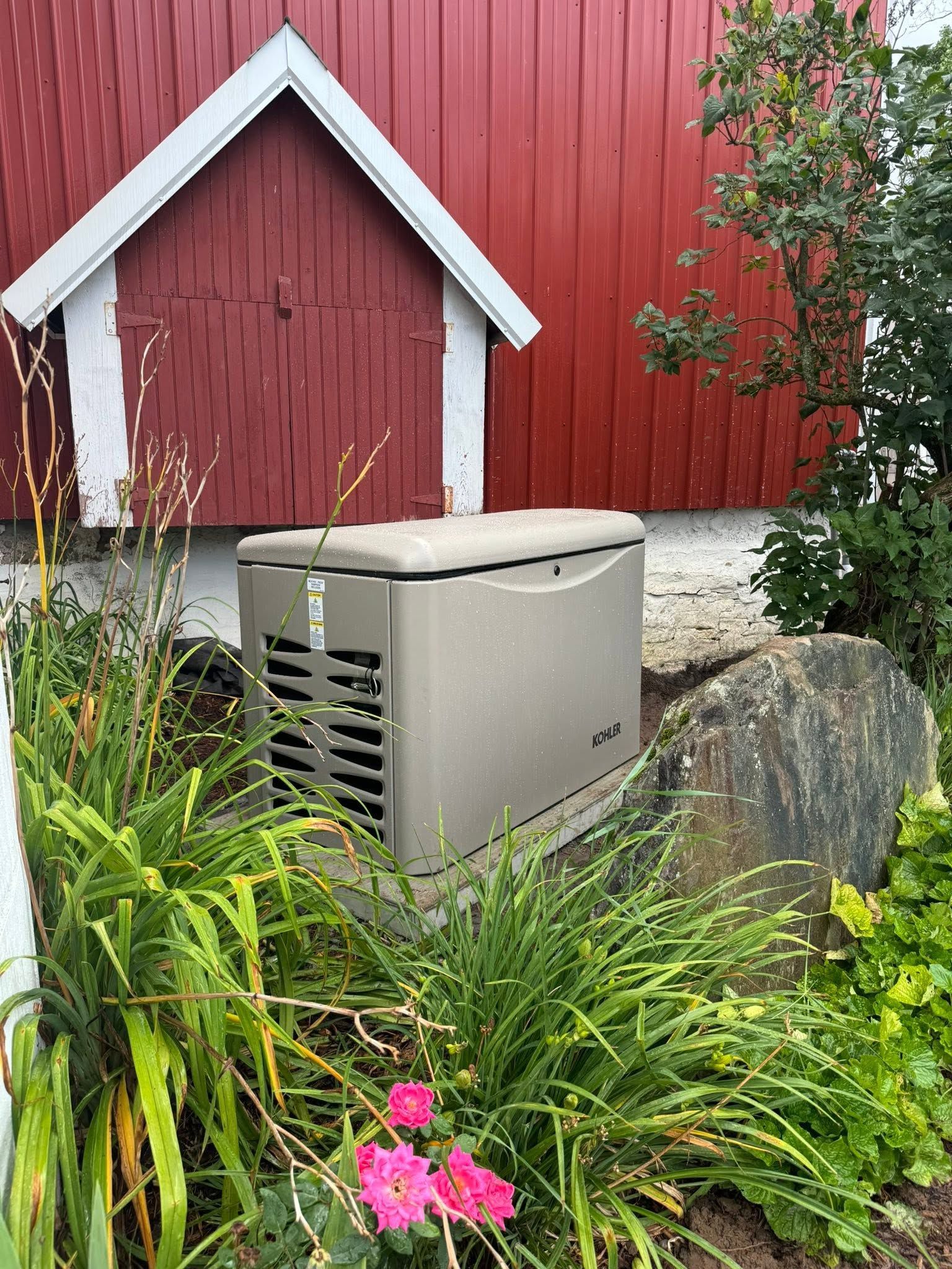 Generator beside red barn wall and garden plants, with flowers and a large rock.