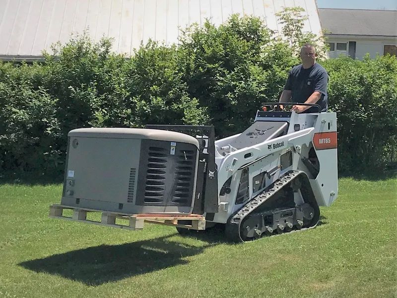 Person driving a compact loader carrying a large outdoor HVAC unit across grass