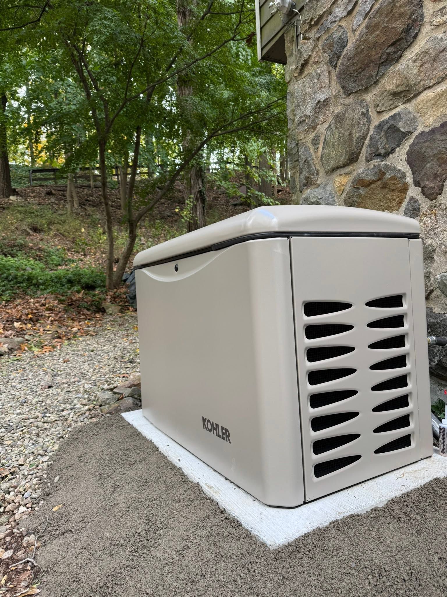 Portable generator on a gravel pad beside a stone wall, with trees and yard in the background