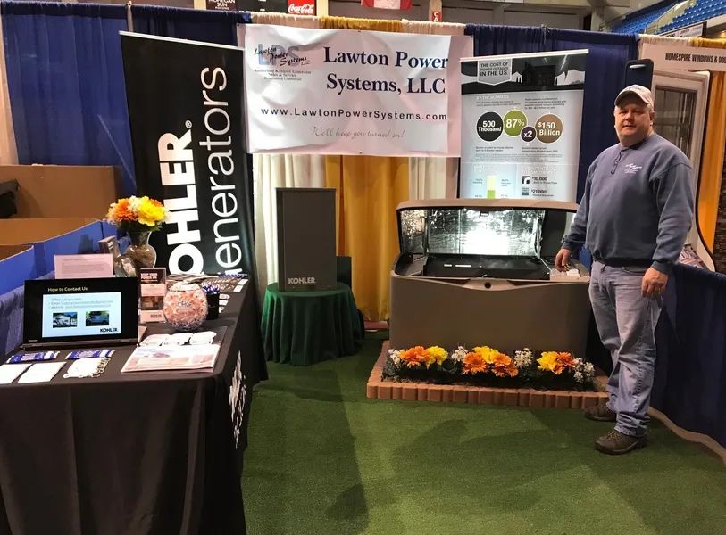 Trade show booth with a man standing beside a large machine, banners, and a monitor on a table.