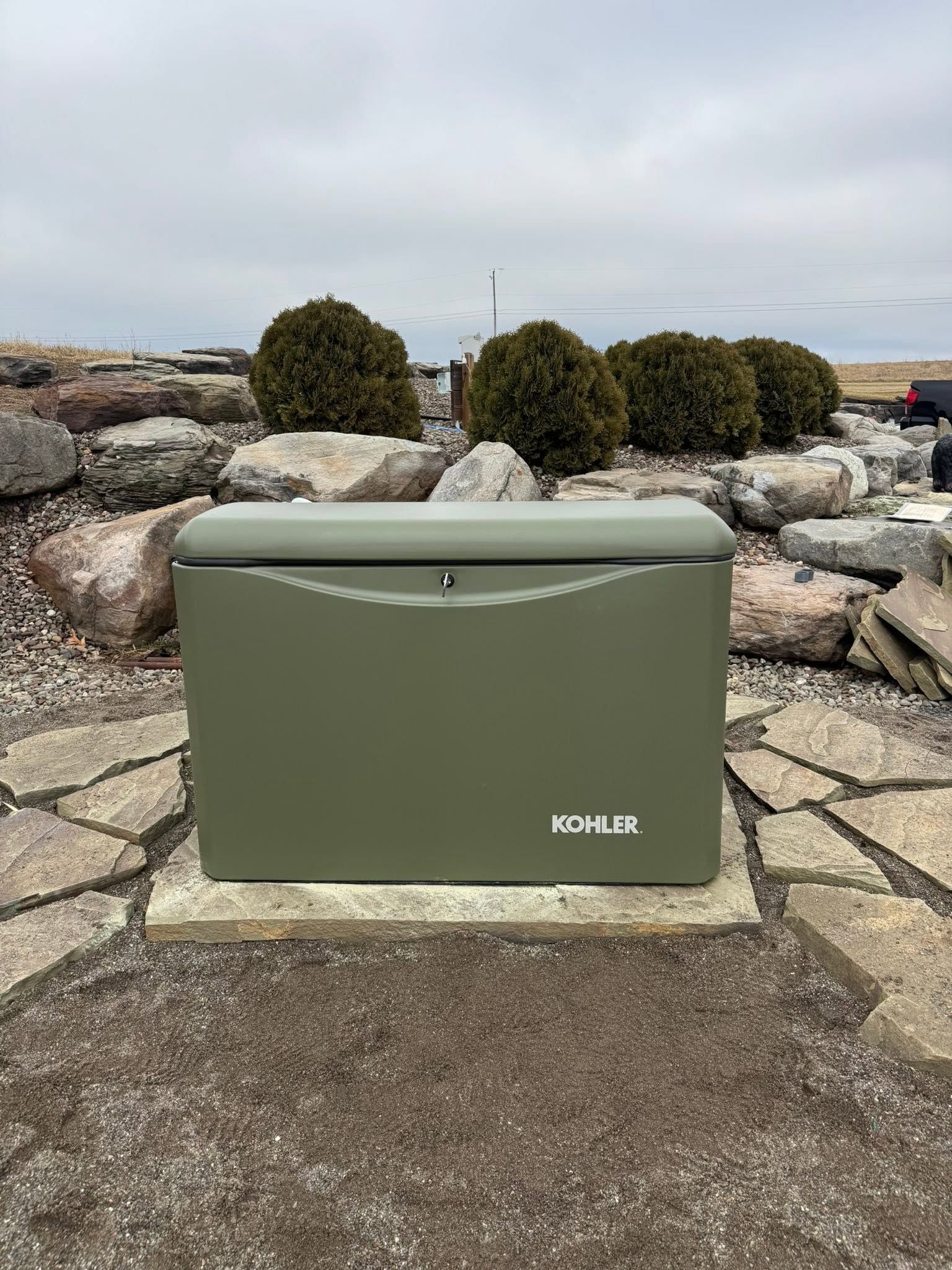 Green MOBIL box on a stone path by rocks and bushes outdoors under a cloudy sky