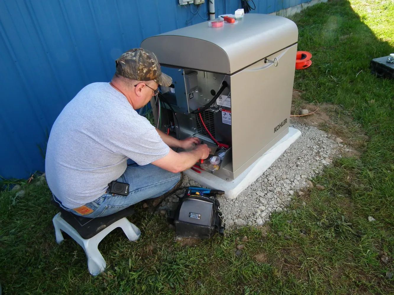 Technician working on an outdoor generator beside a blue building, kneeling on grass and gravel.