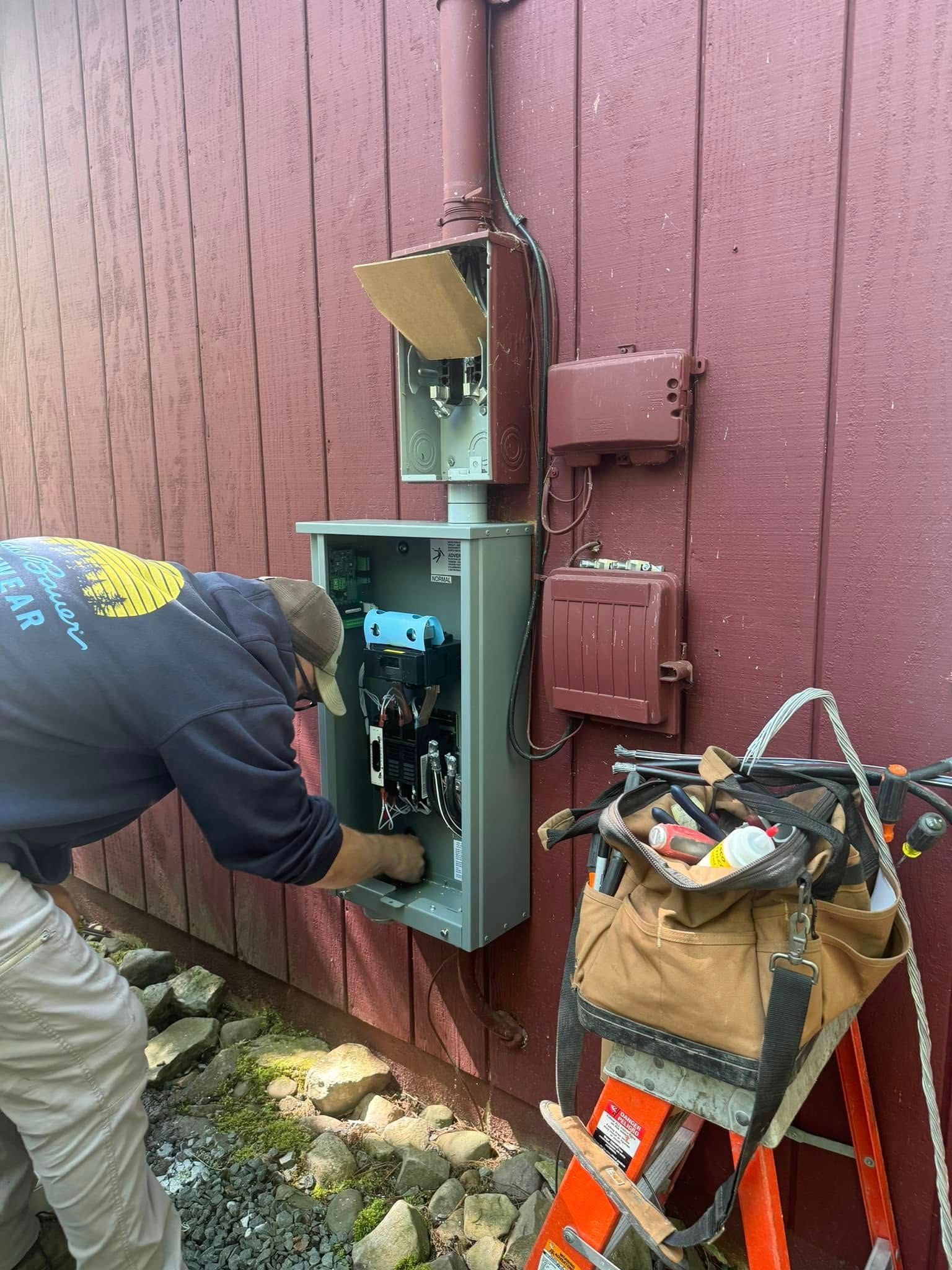 Electrician working on an open electrical panel beside a maroon building, with tools on a ladder nearby.