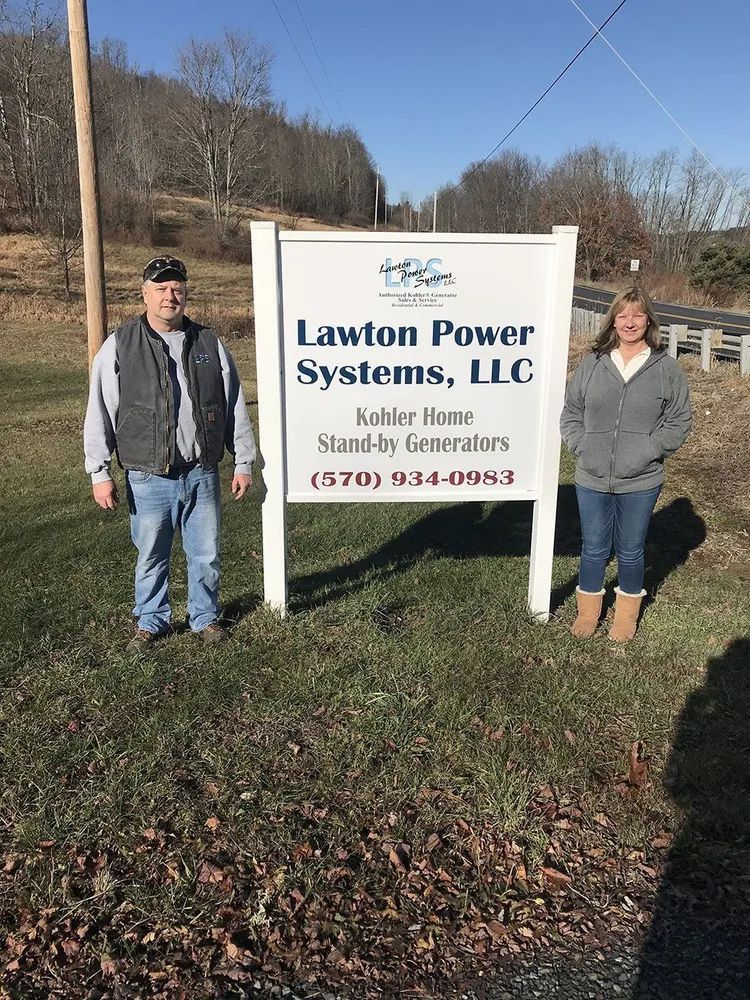 Two people standing beside a Lawton Power Systems, LLC roadside sign on a sunny grassy lot