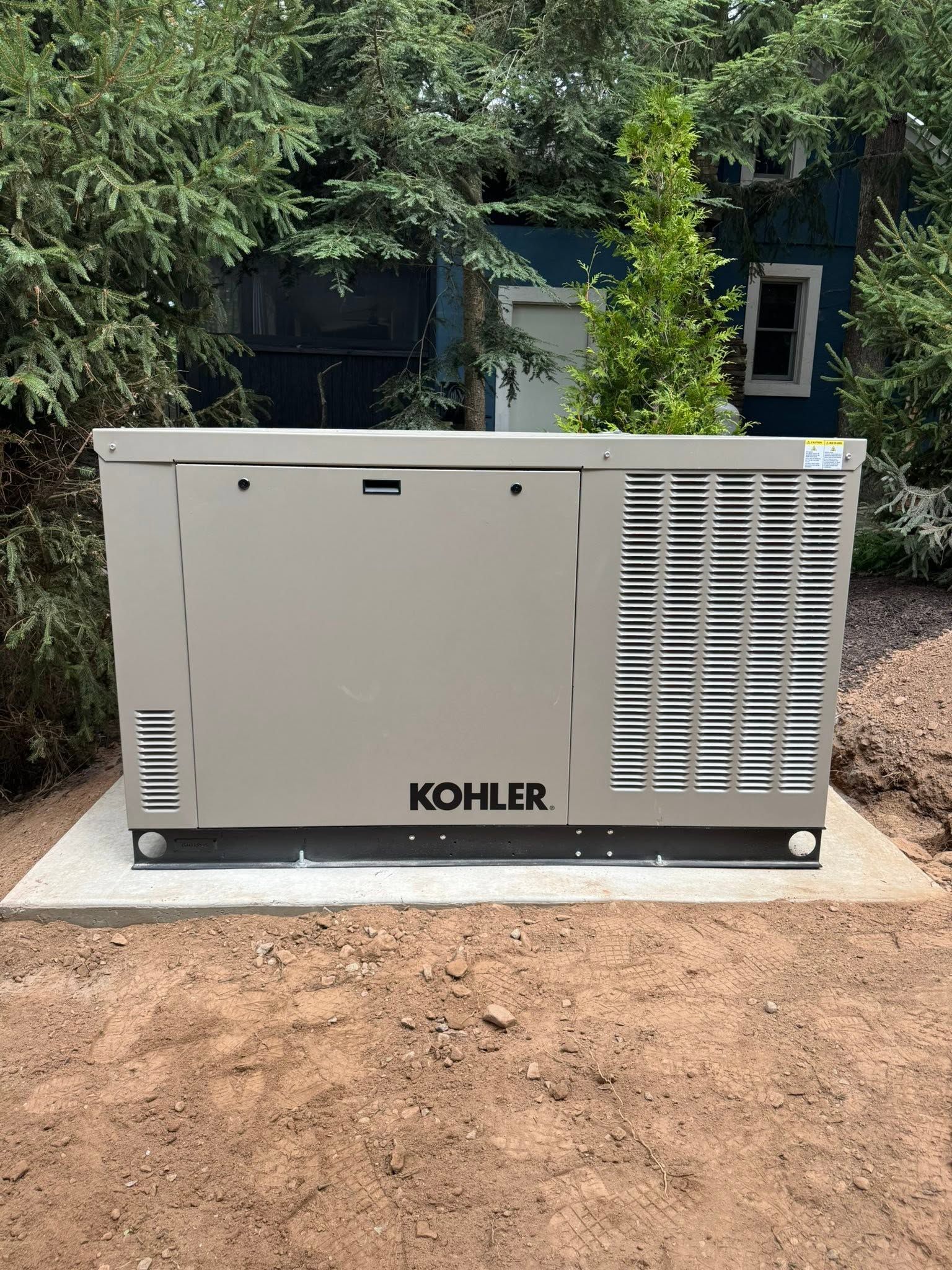 Kohler outdoor generator on a concrete pad beside trees and a dirt yard