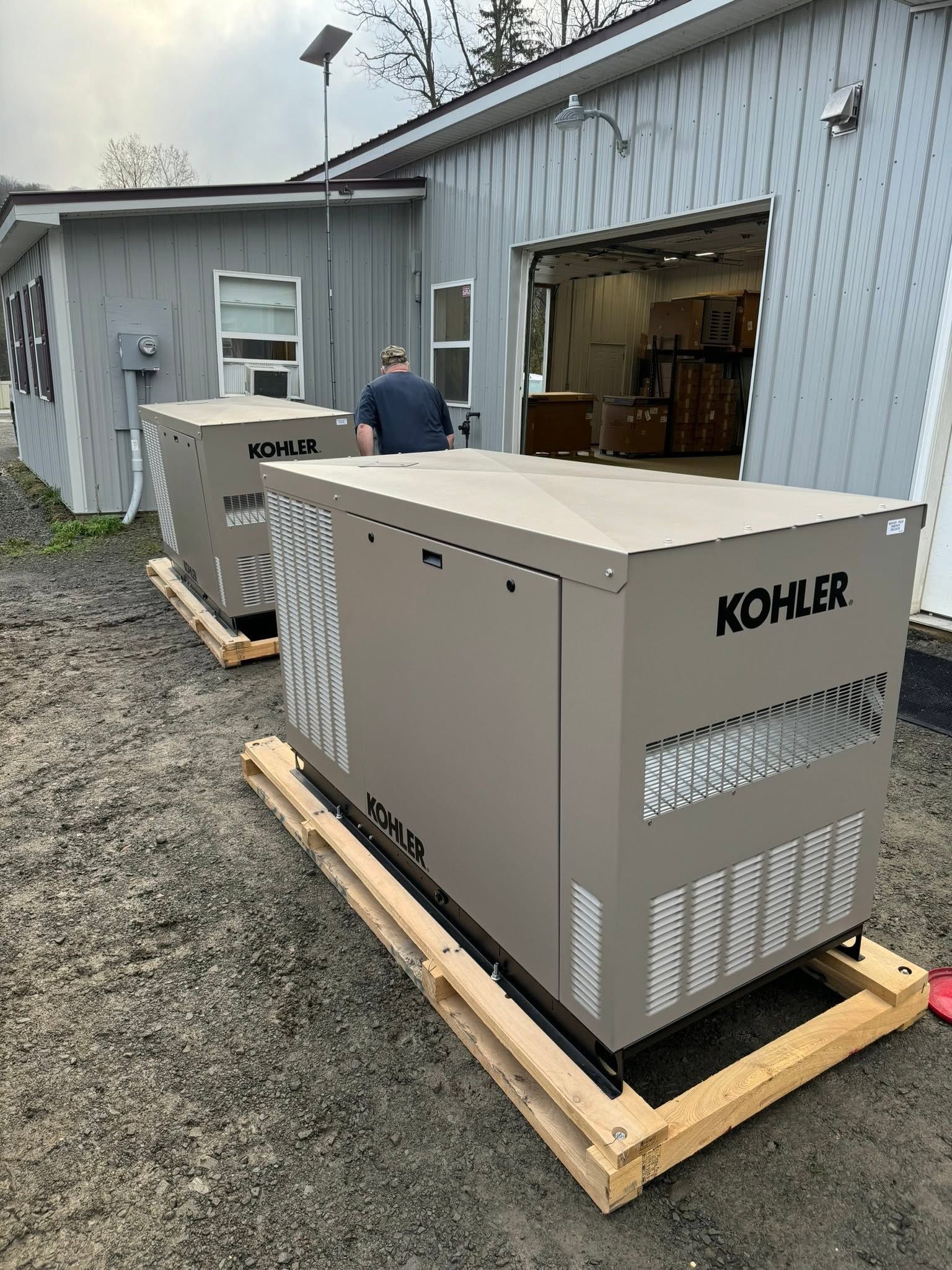 Two Kohler generators on wooden pallets outside a gray building, with a person standing nearby.