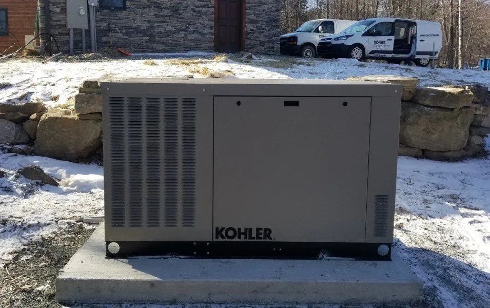 Gray Kohler generator unit on a concrete pad outdoors beside a stone wall and snow.