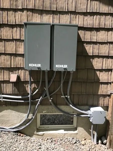 Two Kohler outdoor utility boxes mounted on shingled wall with visible conduit and vent opening below