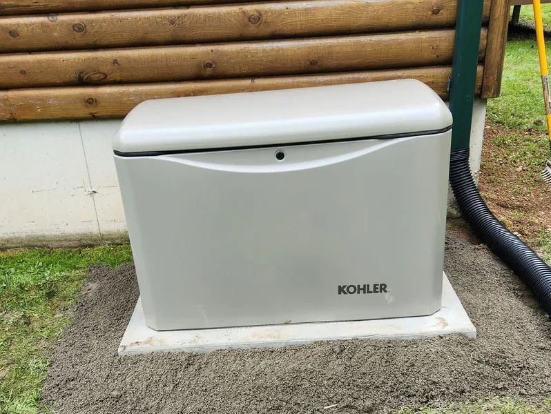 Gray Kohler outdoor storage box on a concrete pad beside a log cabin wall.
