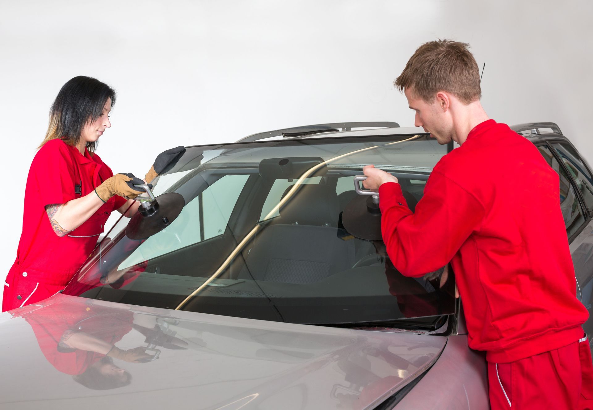 Two technicians in red uniforms install a windshield on a silver car.