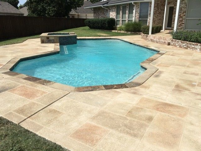Swimming pool with turquoise water surrounded by stone patio. House and grass in background.