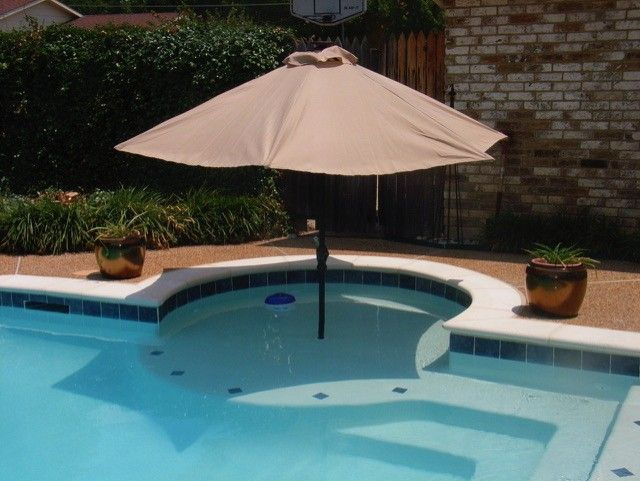 Swimming pool with steps and umbrella, two potted plants on the edge, beige umbrella over the pool.