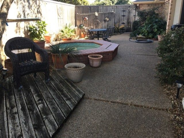Backyard with a hot tub, wooden deck, and patio furniture. Trees and plants are present along the fence.