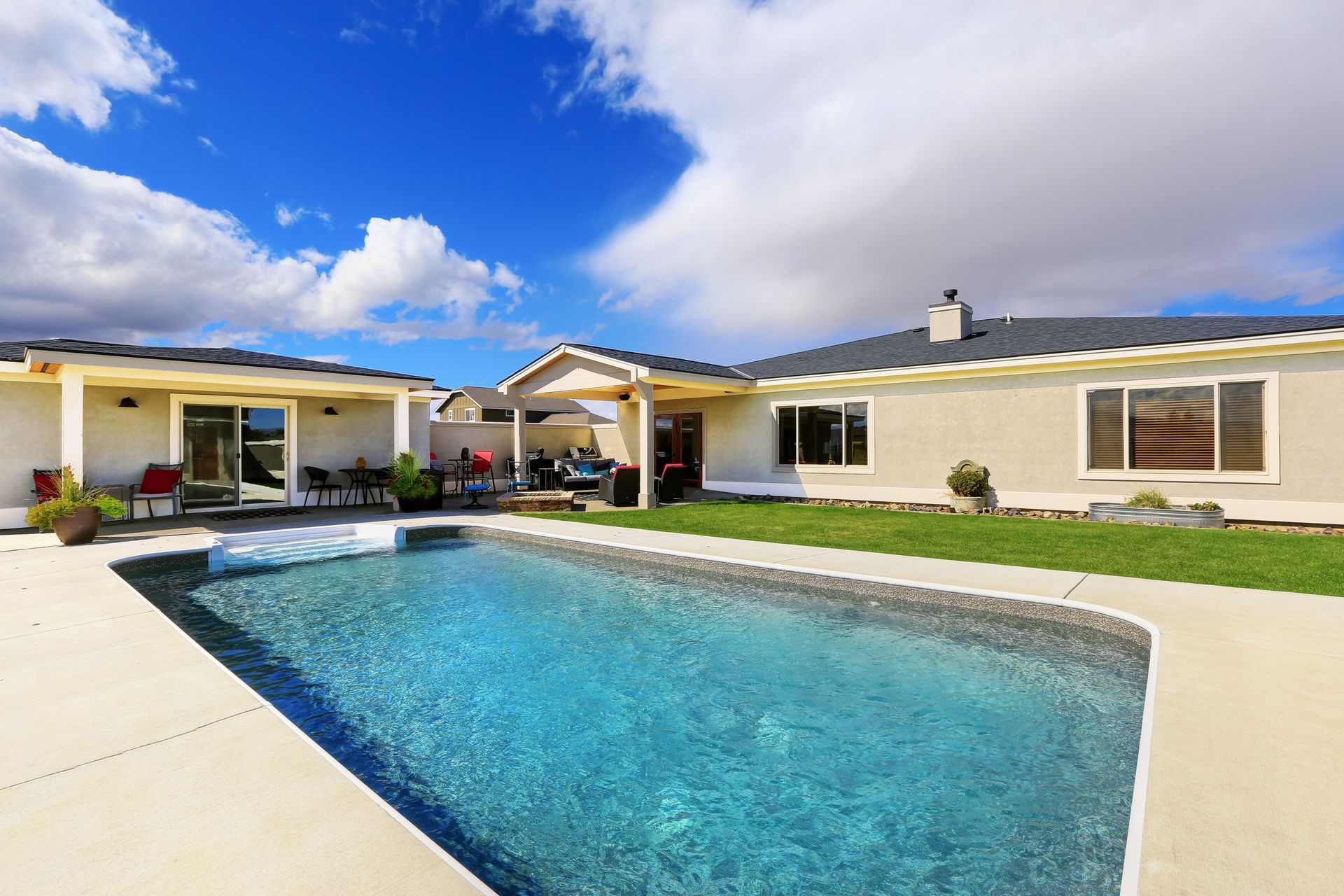 Backyard with a rectangular pool, light-colored house, and blue sky with clouds.