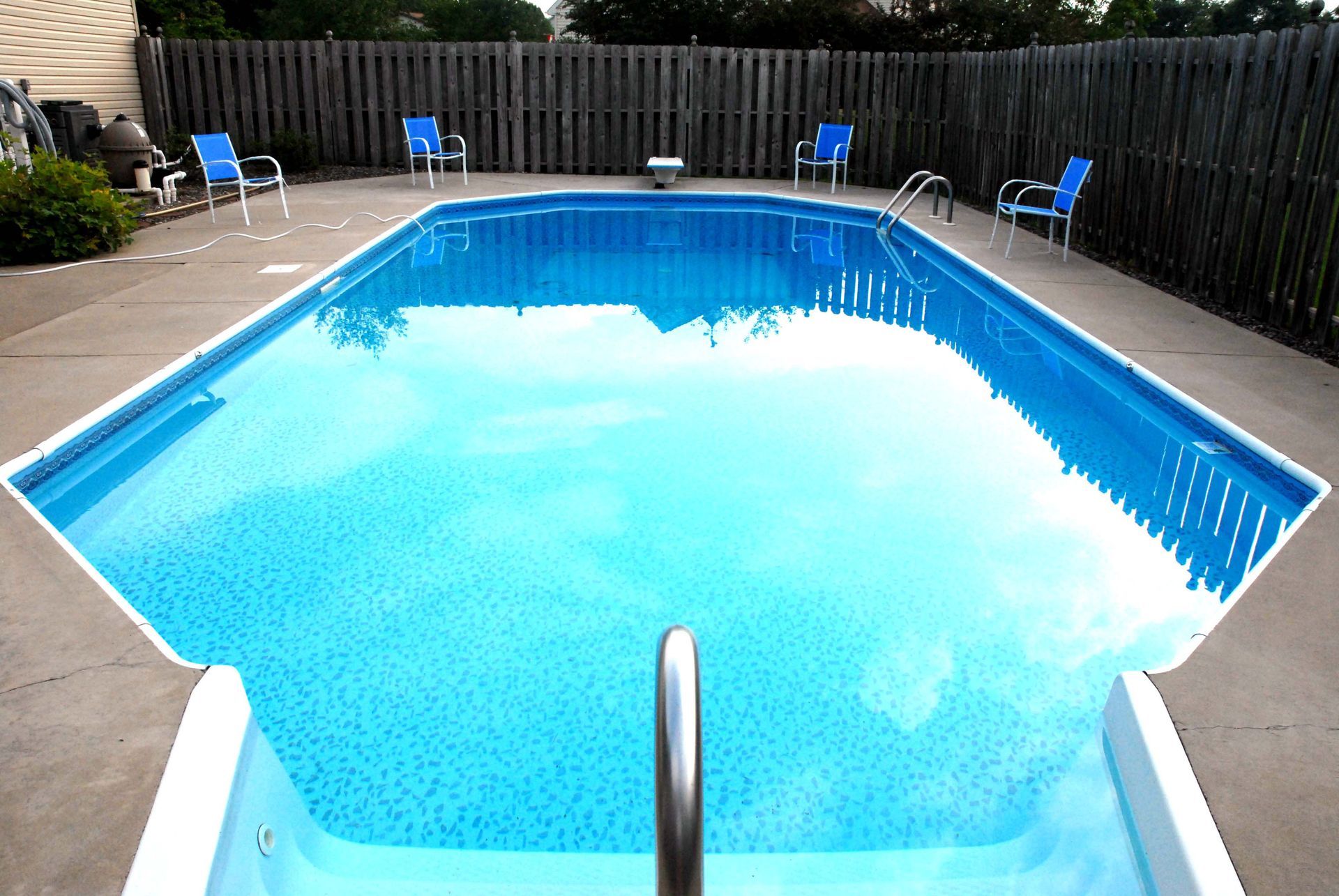 Swimming pool with blue water, surrounded by concrete patio and wooden fence, with blue chairs.