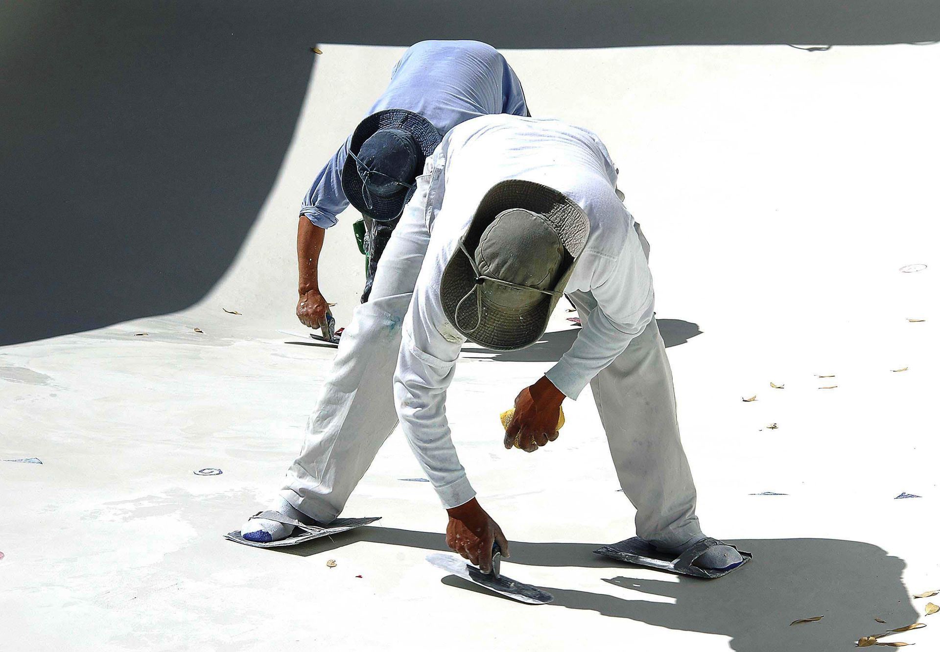 Two workers in a pool, smoothing plaster. One in a blue shirt, the other in white. Bright sunlight.
