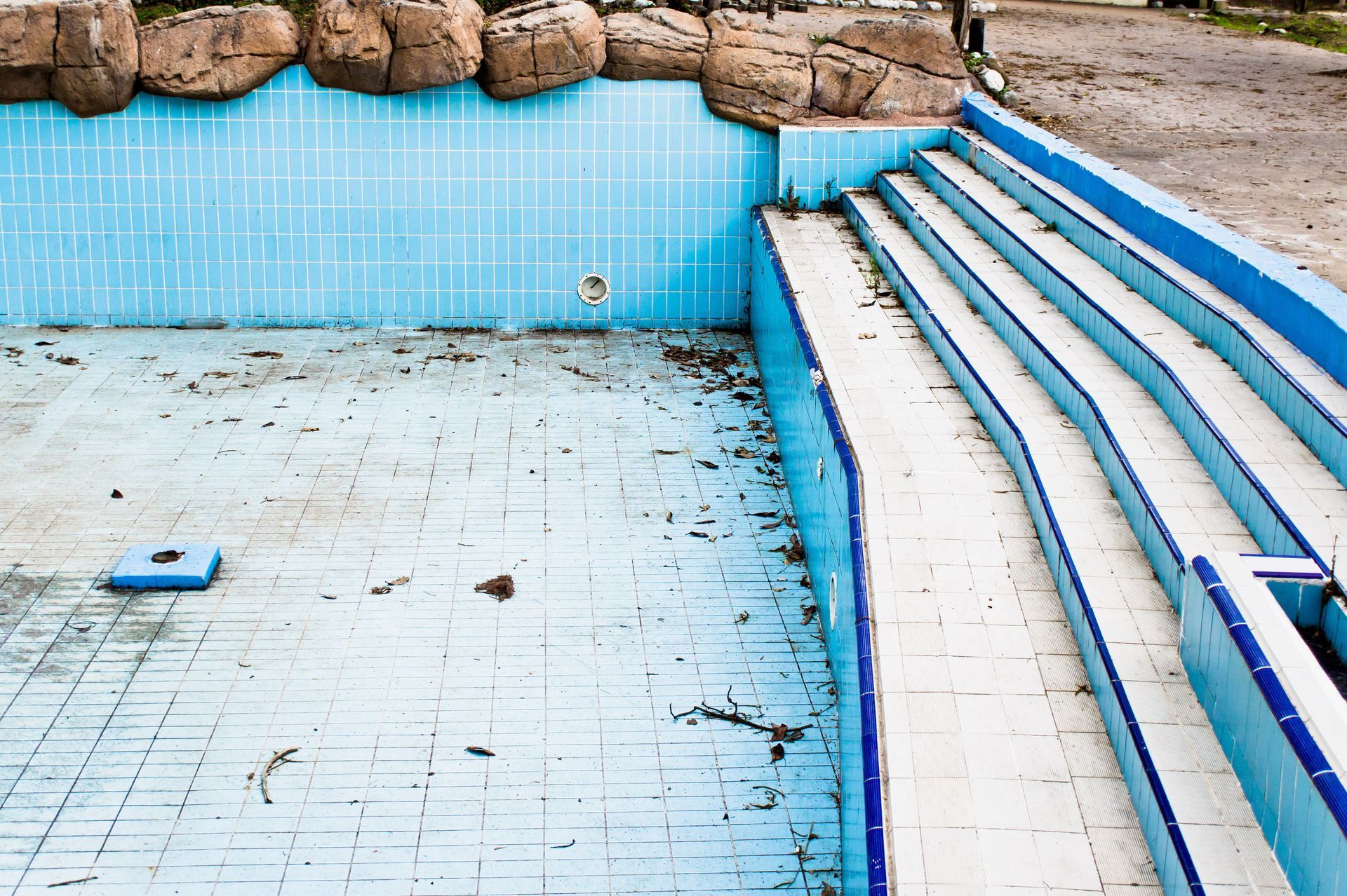 Empty, unmaintained blue tiled pool with steps and rocks.
