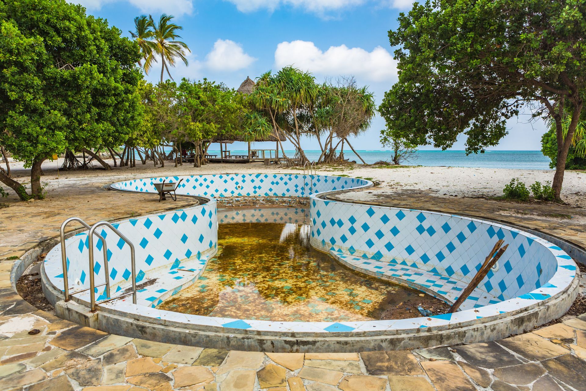Empty, neglected swimming pool with blue and white tile, near a beach with trees.
